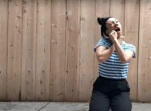 Trixi in a blue stripped shirt and black pants with one hand on her throat and the other miming putting on lipstick.